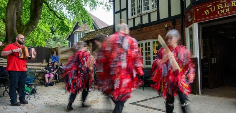 Vibrant red ribbons blur in the ancient rhythm of the dance, a timeless pulse of folklore echoing against the stone walls of The Burley Inn at Beltane.#jerryfennerphotography #newforestphotographer #morrisdancing #beltane #greenmanparade #newforest #folklorerituals #traditionaldance