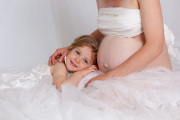 A tender moment captured: a young girl with bright blue eyes rests her head against her mother's pregnant belly, surrounded by soft white fabric. She's listening for the baby and dreaming of becoming a big sister.