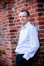 A composed portrait of a man in a crisp blue check shirt, leaning casually against a textured red brick wall. The soft daylight highlights his approachable expression and measured posture, creating an understated, professional mood within this setting.#jerryfennerphotography #portraiture #editorialphotography #professionalportrait #headshots #brickwall #naturaldaylight #menswear #classicstyle #businessportrait #contemporaryphotography #quietmoment